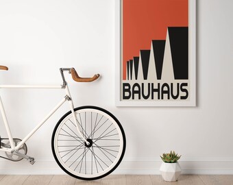 Op de afbeelding: Een Bauhaus-poster met een rode en zwarte geometrische vormgeving hangt in een witte lijst aan een witte muur. Een witte fiets met een bruin leren stuur en een zwarte band staat naast de poster. Een kleine vetplant in een witte geometrische pot staat op de vloer.
