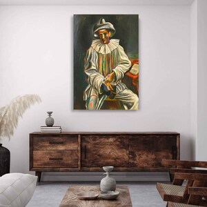 May include: A framed painting of a seated figure in a Pierrot costume. The figure wears a white outfit with colorful stripes, a ruffled collar, and a hat. The background is dark, with a red table and a book visible.