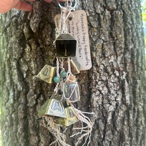 May include: A collection of brass bells, small glass vials, and beads are suspended from a metal ring. The bells are attached with twine and adorned with green beads. A tag reads "Hannah's Happy Handcrafts Lucky Lizard Bells."