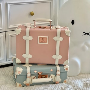 May include: Two vintage-style suitcases with floral designs. The top suitcase is pink with white trim and the bottom suitcase is light blue with white trim. Both suitcases have gold accents and a lock.
