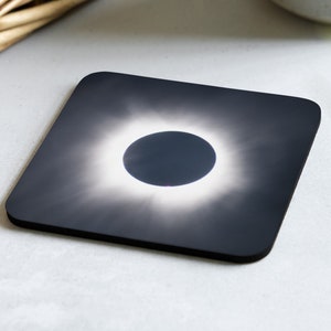 May include: A black square coaster with a photo of a solar eclipse. The sun is a bright white ring around a dark circle.