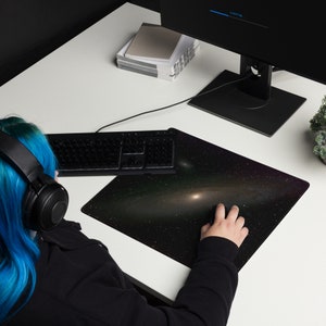 May include: A rectangular mousepad featuring a galaxy design with a central bright nebula and surrounding stars. The mousepad is black with a colorful cosmic pattern. A black keyboard and mouse are on the white desk.