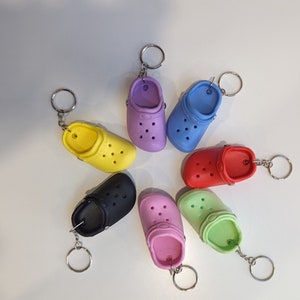 May include: Seven colourful plastic clog-shaped keychains. The keychains are in the colours purple, blue, yellow, red, black, pink, and green.