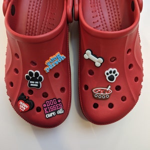 May include: A red pair of clogs with various dog-themed Jibbitz charms. Charms include "Dog Person", "Dog on Board", "Puppy Love", "Dog Kisses Cure All", a bone, a paw print, and a bowl of chilli.