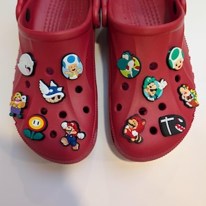 May include: A pair of red clogs decorated with cartoon character charms. The charms depict characters from the Super Mario Bros. series, such as Mario, Luigi, and Yoshi. The clogs are made of a rubbery material and have several ventilation holes.