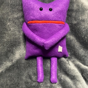 May include: A purple felt plush toy shaped like a creature with two small black bead eyes, a straight red horizontal line for a mouth, two arms folded across the front, and two dangling legs. A small white square patch is attached to the lower right.