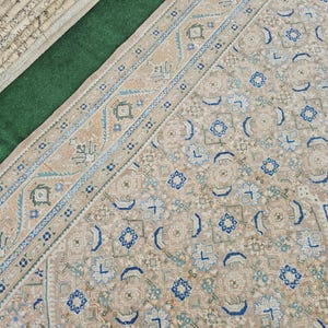 Puede incluir: Vista a&eacute;rea de una alfombra estampada en tonos beige y azul. La alfombra tiene un borde detallado con dise&ntilde;os geom&eacute;tricos y florales. La paleta de colores incluye beige, azul y verde. Alfombras enrolladas son visibles en la esquina superior izquierda.