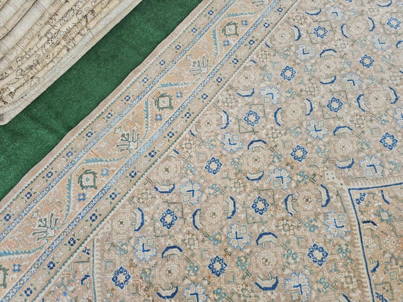 Puede incluir: Vista a&eacute;rea de una alfombra estampada en tonos beige y azul. La alfombra tiene un borde detallado con dise&ntilde;os geom&eacute;tricos y florales. La paleta de colores incluye beige, azul y verde. Alfombras enrolladas son visibles en la esquina superior izquierda.