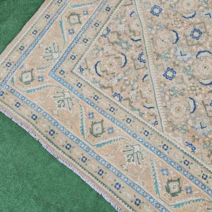 Puede incluir: Una alfombra beige y azul estampada con un borde detallado. La alfombra presenta un dise&ntilde;o central con formas geom&eacute;tricas y motivos florales en tonos beige, azul y verde. El borde es azul oscuro con patrones intrincados.