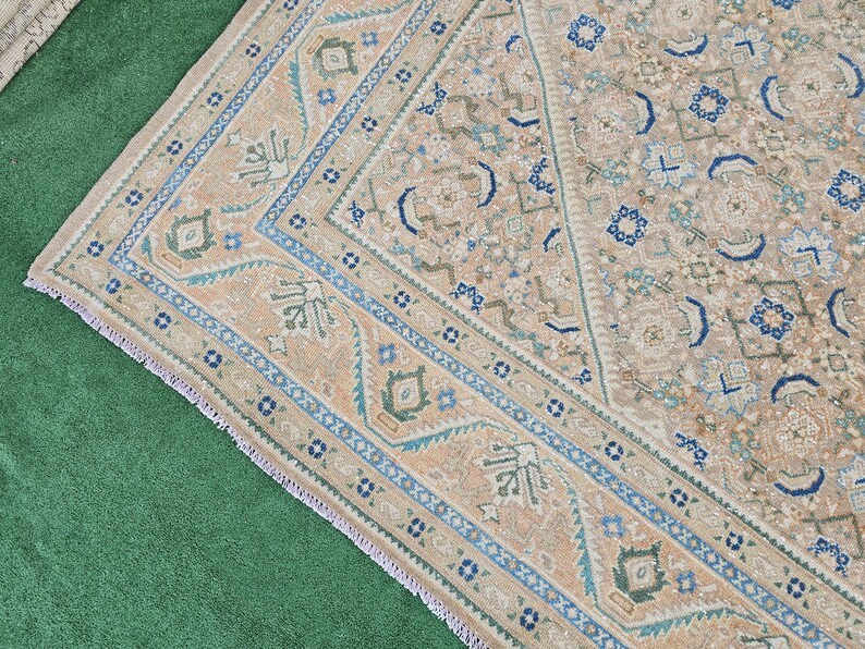 Puede incluir: Una alfombra beige y azul estampada con un borde detallado. La alfombra presenta un dise&ntilde;o central con formas geom&eacute;tricas y motivos florales en tonos beige, azul y verde. El borde es azul oscuro con patrones intrincados.