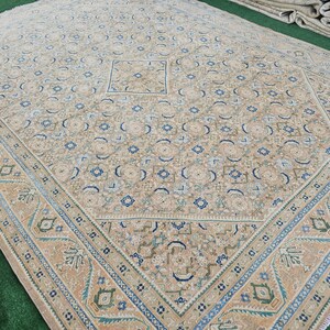 Puede incluir: Gran alfombra beige con un intrincado patr&oacute;n geom&eacute;trico. La alfombra presenta un dise&ntilde;o cuadrado central y un borde con detalles en azul, verde y beige. El dise&ntilde;o general es sim&eacute;trico y detallado, adecuado para una sala de estar o comedor.
