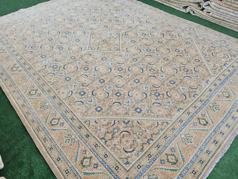Puede incluir: Gran alfombra beige con un intrincado patr&oacute;n geom&eacute;trico. La alfombra presenta un dise&ntilde;o cuadrado central y un borde con detalles en azul, verde y beige. El dise&ntilde;o general es sim&eacute;trico y detallado, adecuado para una sala de estar o comedor.