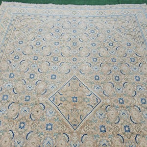 Puede incluir: Una alfombra rectangular grande con un patr&oacute;n sim&eacute;trico intrincado. La alfombra presenta un dise&ntilde;o de diamante central y motivos florales repetidos en tonos beige, azul y verde. El borde de la alfombra es de un tono beige m&aacute;s claro con patrones geom&eacute;tricos.
