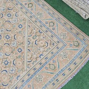 Puede incluir: Una secci&oacute;n de una alfombra beige y azul estampada con un borde detallado. La alfombra presenta un dise&ntilde;o central con elementos florales y geom&eacute;tricos en tonos azules, beige y verdes. La alfombra est&aacute; sobre una superficie verde.