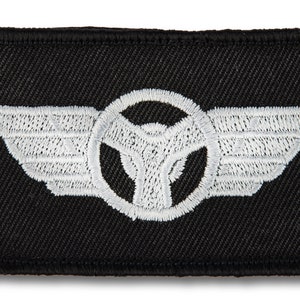 May include: White embroidered patch on a black background. The patch features a stylised steering wheel with wings on either side.