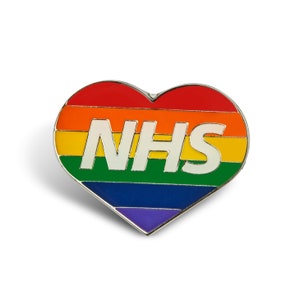 May include: A heart-shaped enamel pin featuring the NHS logo. The pin has a rainbow design with the letters "NHS" in white. The colours are red, orange, yellow, green, blue, and purple, with a silver border and white background.