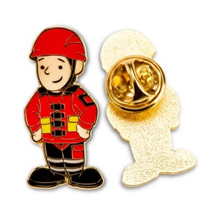 May include: Gold-coloured enamel pin featuring a cartoon firefighter wearing a red helmet and a yellow and black uniform. The pin has a safety clasp on the back.