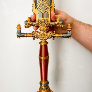 May include: A gold and red decorative sword with intricate details. The hilt is shaped like a castle with a pointed top and a window in the center. The handle is red with gold accents. The sword is held by a hand.