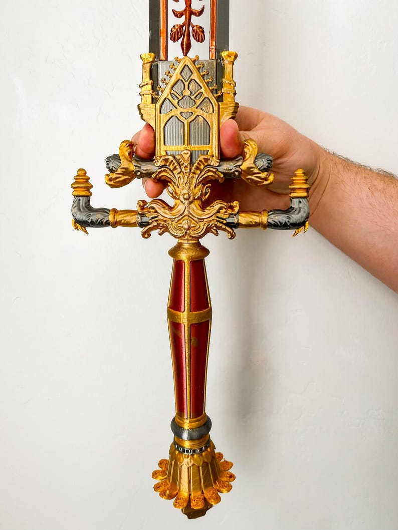 May include: A gold and red decorative sword with intricate details. The hilt is shaped like a castle with a pointed top and a window in the center. The handle is red with gold accents. The sword is held by a hand.