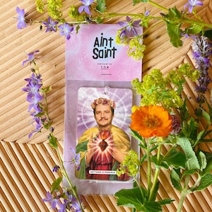 Peut inclure: Un désodorisant avec un portrait d'homme portant une couronne de fleurs et une cape dorée, avec le texte « Anything is Possible ». Le désodorisant est dans un emballage plastique transparent avec la marque « Aint Saint » sur fond rose. L'image est entourée de fleurs.