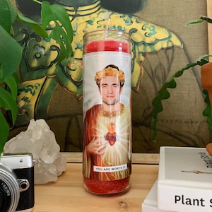 May include: Red candle with a white label featuring a portrait of a man with a crown of flowers and the text "You are worth it".