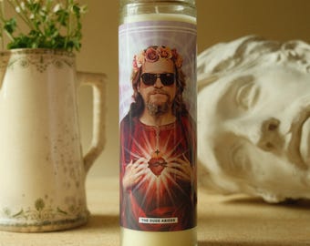 May include: A glass candle with a label featuring a portrait of a man wearing sunglasses and a flower crown. The man's image is superimposed over a red and yellow burst design. The text "THE DUDE ABIDES" is at the bottom of the label.