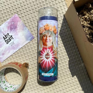 May include: A blue and white candle featuring a picture of a woman with curly hair wearing a flower crown. The candle has the text "Enjoy Yourself" at the bottom.