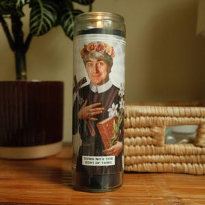 May include: A glass candle with a printed image of a person wearing a floral crown and holding a book. The text on the candle reads "Down with this sort of thing."