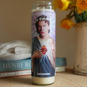 May include: A glass candle with a printed image of a person wearing a floral crown and holding a heart. The candle has the text "NO ONE ELSE WILL DO" at the bottom. The candle is on a wooden surface.
