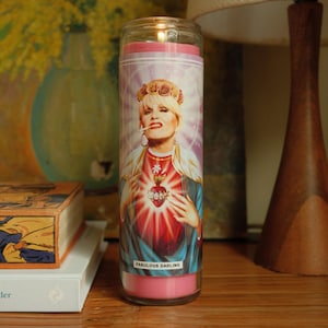 May include: A pink candle featuring a picture of a woman with blonde hair wearing a blue and red outfit. The woman is holding a red heart with a floral design. The text "FABULOUS DARLING" is printed on the candle.