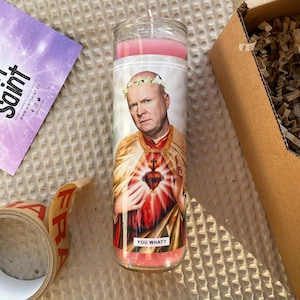 Op de afbeelding: Een roze kaars met een afbeelding van een man met een bloemenkrans op zijn hoofd. De man draagt een rode en gouden mantel en heeft een hart met een kruis op zijn borst. De tekst "YOU WHAT?" staat op de onderkant van de kaars.