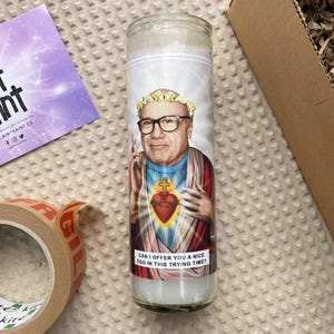 May include: A white candle with a colorful image of a man wearing a crown of flowers and a blue and red robe. The text on the candle reads "Can I offer you a nice egg in this trying time?"