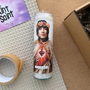 Pode incluir: Uma vela branca com uma imagem impressa de uma pessoa usando uma coroa de flores e uma t&uacute;nica. A pessoa est&aacute; segurando um cora&ccedil;&atilde;o com uma cruz nele. O texto "That's the Spirit" est&aacute; impresso abaixo da imagem.