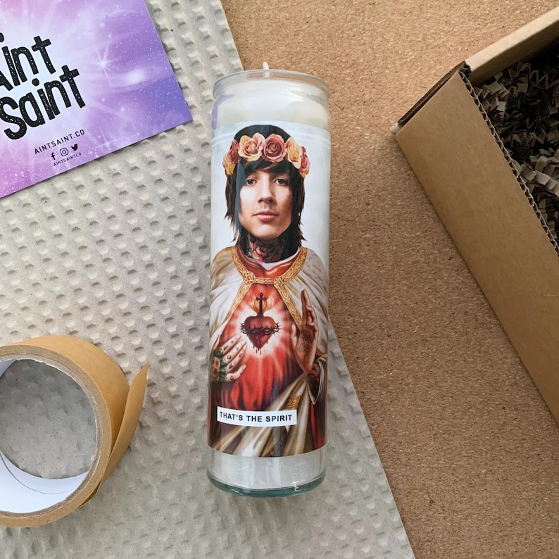 Pode incluir: Uma vela branca com uma imagem impressa de uma pessoa usando uma coroa de flores e uma t&uacute;nica. A pessoa est&aacute; segurando um cora&ccedil;&atilde;o com uma cruz nele. O texto "That's the Spirit" est&aacute; impresso abaixo da imagem.