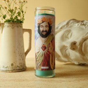 May include: A glass candle featuring a portrait of a man with a floral crown and a heart. The candle has a teal base and a green wick. The words "HAVE FAITH" are printed on the candle.