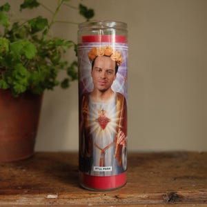 May include: A tall cylindrical red wax candle in a clear glass container featuring a label with a portrait of a person wearing a gold floral crown, depicted in a religious style with a glowing heart and the text IT'LL PASS printed at the bottom.