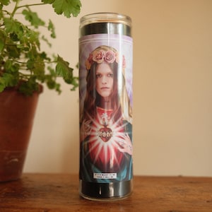 May include: A cylindrical glass prayer candle featuring a graphic label of a person wearing a floral crown and a veil. The person has a glowing heart on their chest with radiating light. The text at the bottom reads, IF IT'S MEANT TO BE THEN IT WILL BE.