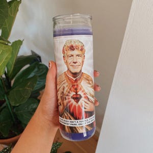 May include: A purple glass candle with a printed image of a man wearing a flower crown. The text on the candle reads "Your body isn't a temple, it's an amusement park. Enjoy the ride."