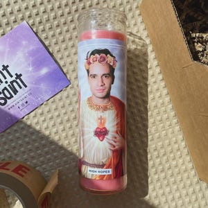 May include: A tall, pink-topped candle with a printed image of a person wearing a floral crown and religious garb. The candle has the words "HIGH HOPES" at the bottom. A purple card with text and a cardboard box are in the background.