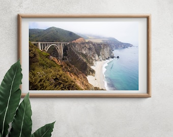 May include: A scenic photograph of a concrete bridge over a coastal highway with a view of a sandy beach and the ocean. The bridge is surrounded by lush green vegetation and the ocean is a vibrant blue.