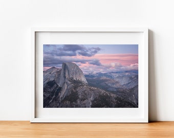 Może przedstawiać: Panoramiczny widok na Half Dome, granitową kopułę w Parku Narodowym Yosemite w Kalifornii. Niebo jest w żywych odcieniach różu i fioletu z puszystymi chmurami. Góra jest otoczona bujną zielenią i doliną.