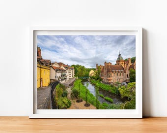 May include: A picturesque view of a river winding through a historic town. The river is bordered by verdant greenery and trees. On one side of the river, there are several buildings with yellow and red brick facades. On the other side, a large stone building with a tower stands tall. The sky is a mix of blue and white clouds.