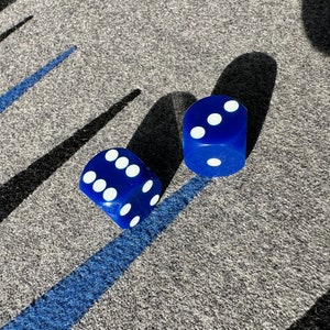 Puede incluir: Dos dados azules con puntos blancos sobre una superficie gris. Los dados están en foco y la superficie está desenfocada.