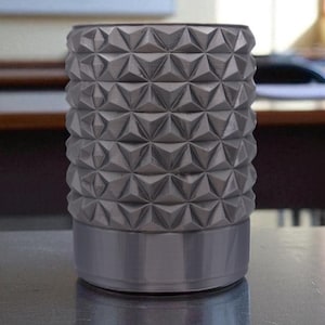 May include: A cylindrical, silver-colored container with a geometric, diamond-like pattern. The container sits on a reflective surface, with a classroom setting in the background. The design features a series of repeating, three-dimensional shapes.
