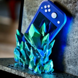 May include: A blue Nintendo Switch Lite video game console rests on a 3D printed stand made of iridescent green and blue crystals.