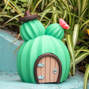 May include: A whimsical, green cactus-shaped house with a brown sombrero on top. The house features a wooden door with a brown frame and blue accents. A pink flower adorns one of the cactus arms. The house is set against a backdrop of green foliage.