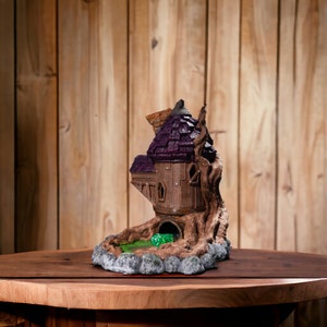 May include: A 3D printed miniature treehouse with a purple roof and a brown wooden base. The treehouse is set on a brown base with a green pond and rocks.