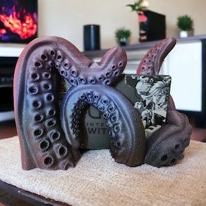 May include: A 3D-printed octopus tentacle-shaped stand, in shades of purple and brown, designed to hold a Nintendo Switch console. The stand features detailed suckers and a textured surface, with the Nintendo Switch logo visible.