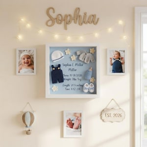 May include: A white shadow box frame containing a baby hat, a cardigan, a pair of booties, a bottle, and a navy blue romper. The text reads: Sophia E. Miller Miller, Born: 11/18/2026, Weight 7lbs 9oz, Length: 20.5 inches, Time: 8:12 AM. Surrounded by photos.