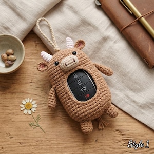 May include: A brown crocheted cow-shaped key fob cover featuring a light tan snout, small pink-tipped horns, and black bead eyes. The center opening reveals a black car key fob with four buttons. A braided cord loop is attached to the top.